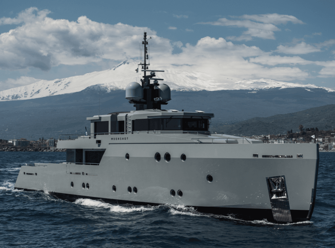 A sleek yacht named "Moonshot" sails near a coastal town, with a snow-capped mountain and cloudy sky in the background.