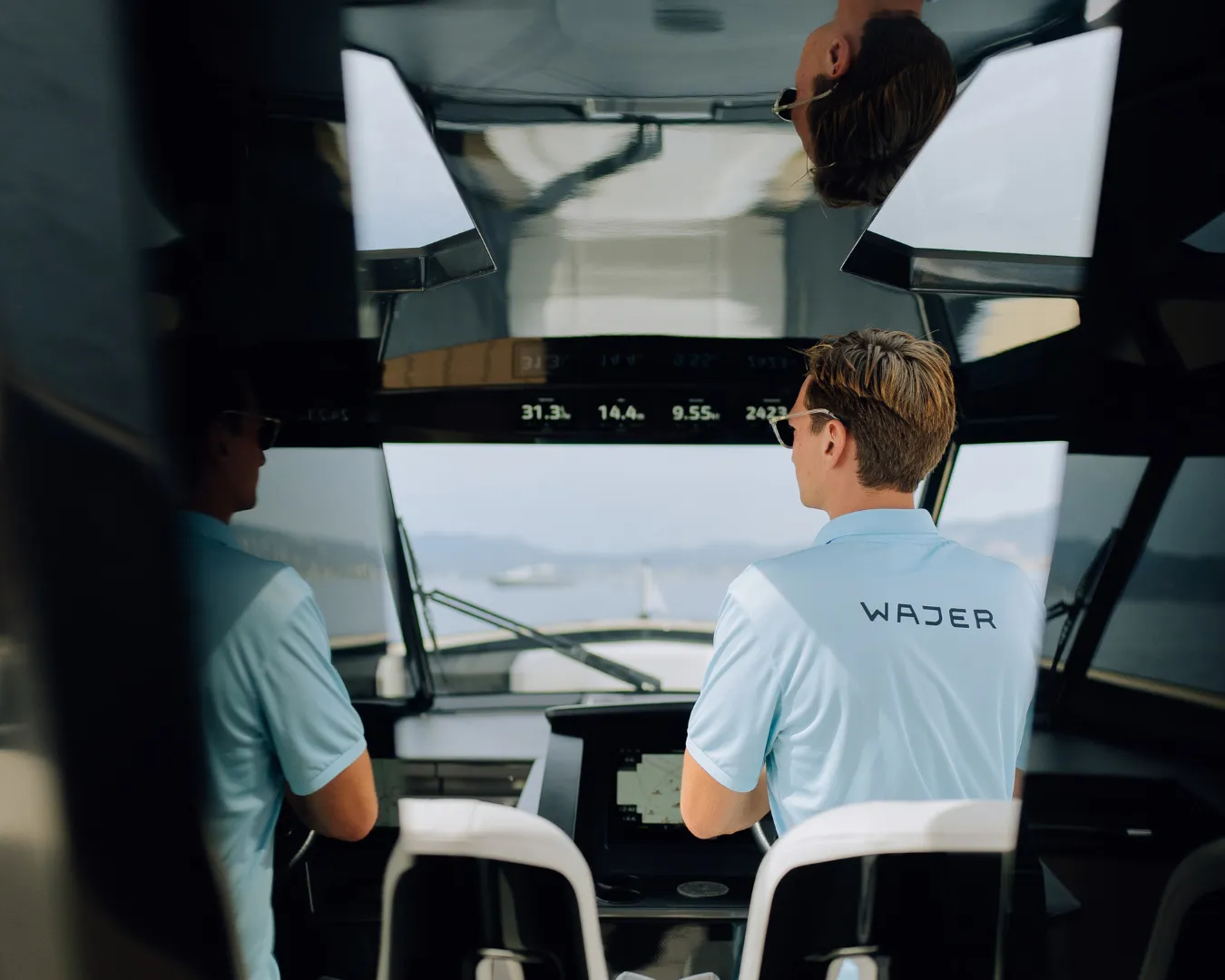 Person in a blue shirt with "Wajer" logo, navigating a boat, with a reflection on the ceiling and distant mountains visible through the window.