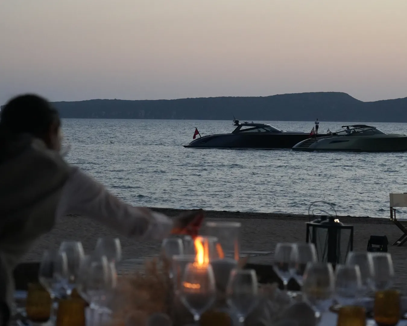 Boats anchored near the shore at sunset with a blurred person lighting a candle in the foreground and a set table with glasses.