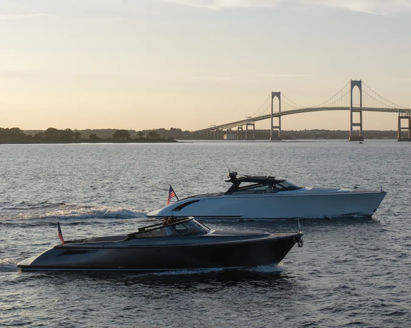 Two motorboats glide on the water near a large suspension bridge at sunset, with American flags on their sterns.