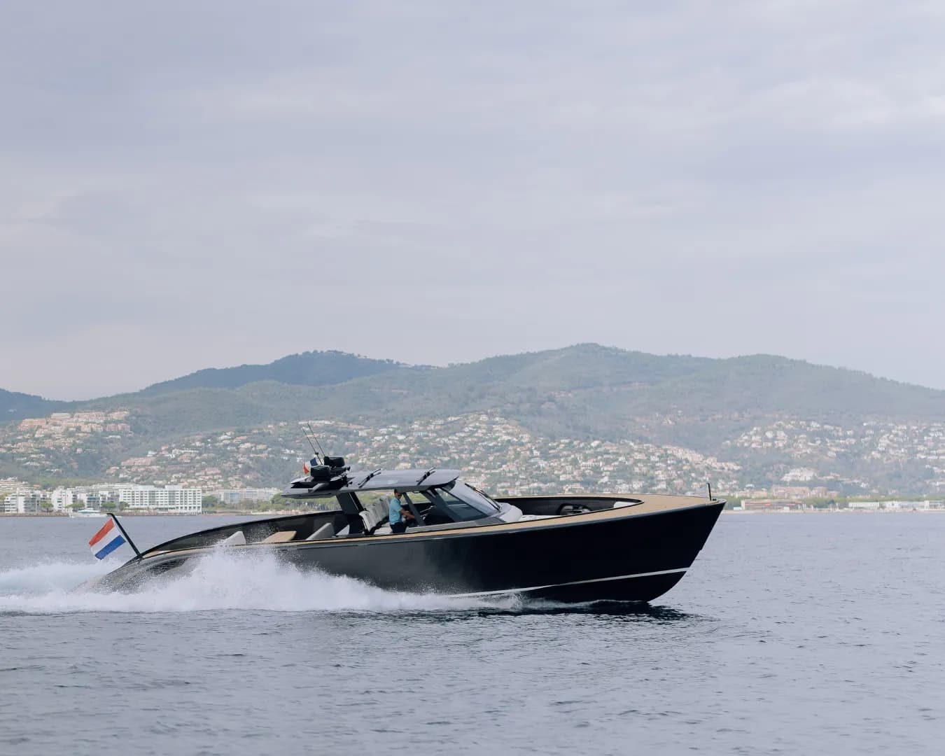 A sleek black motorboat speeds across the water, displaying a red, white, and blue flag, with a scenic backdrop of hills and coastal buildings.