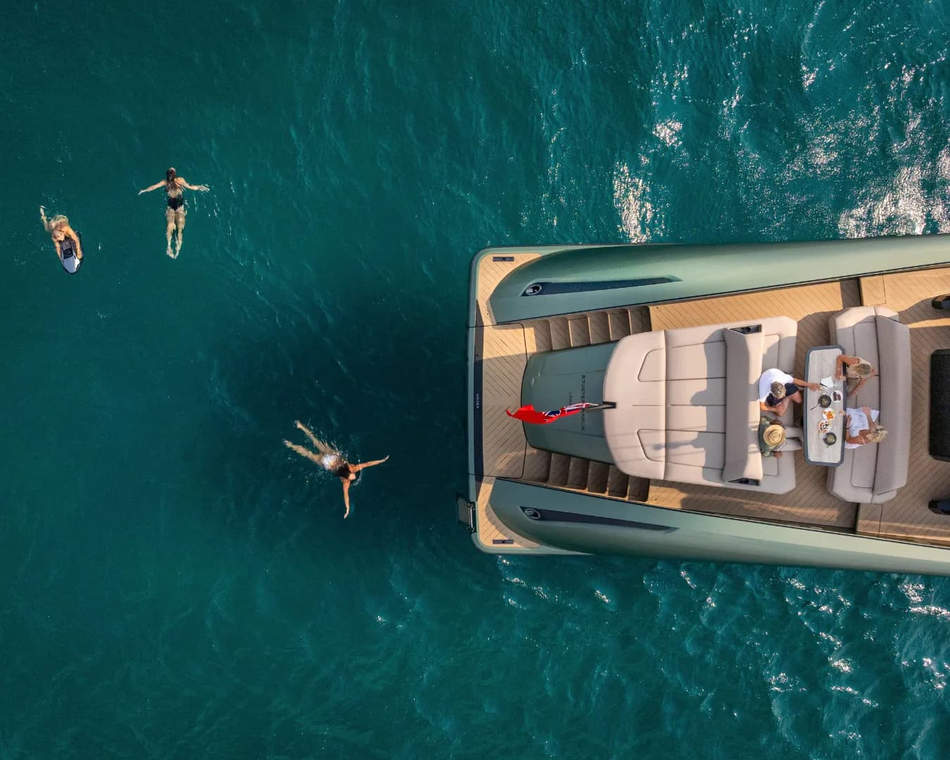 Aerial view of people swimming near a luxurious yacht in clear blue water, with others seated on the yacht's deck enjoying a meal.