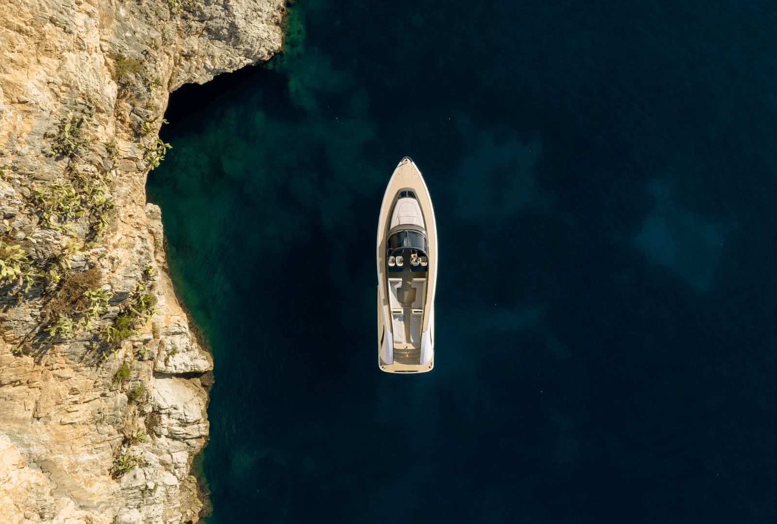 Aerial view of a yacht near a rocky coastline with deep blue water, highlighting the contrast between the natural and man-made elements.