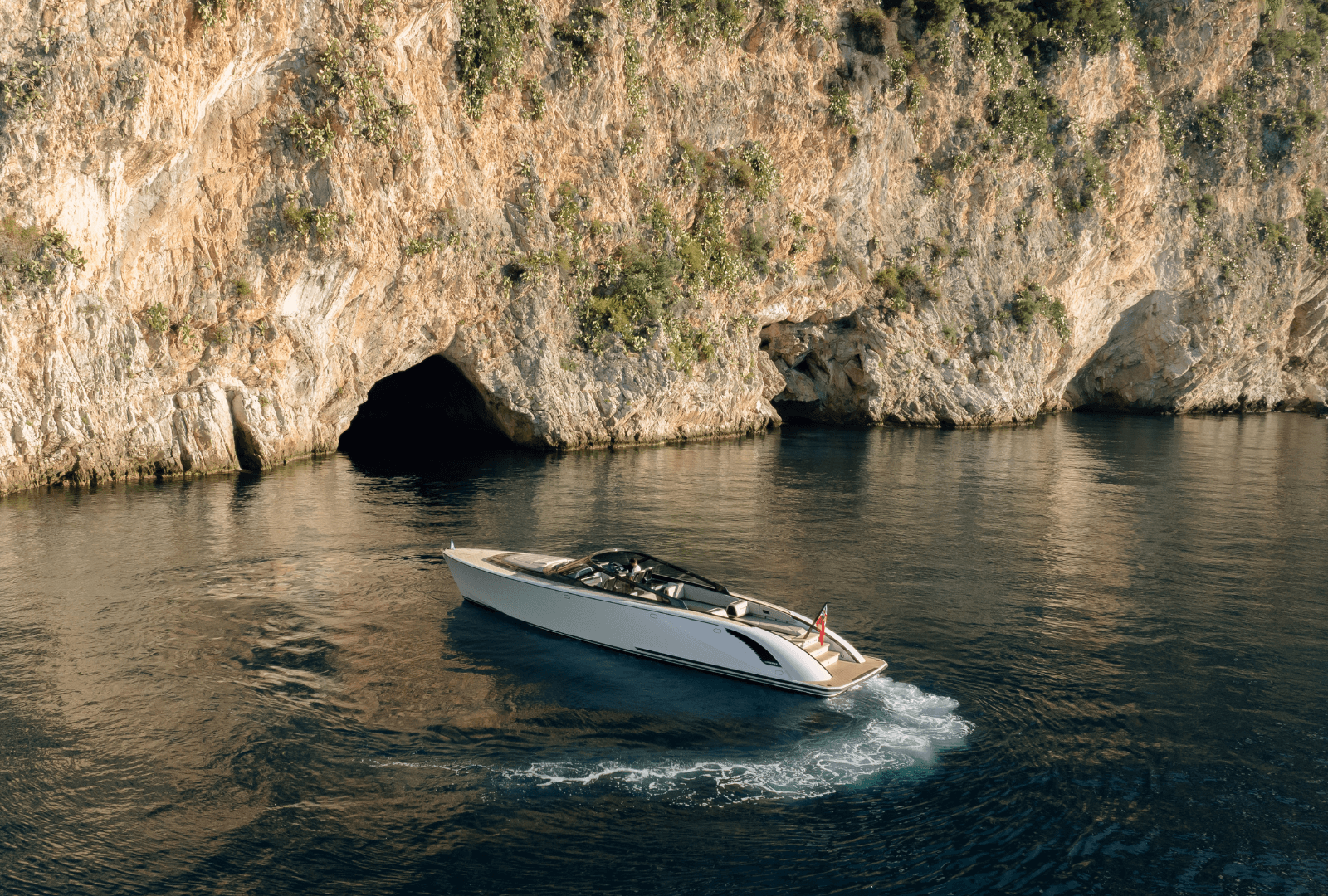 A sleek boat cruising near rocky cliffs, with a small cave visible in the background and calm, reflective water surrounding it.