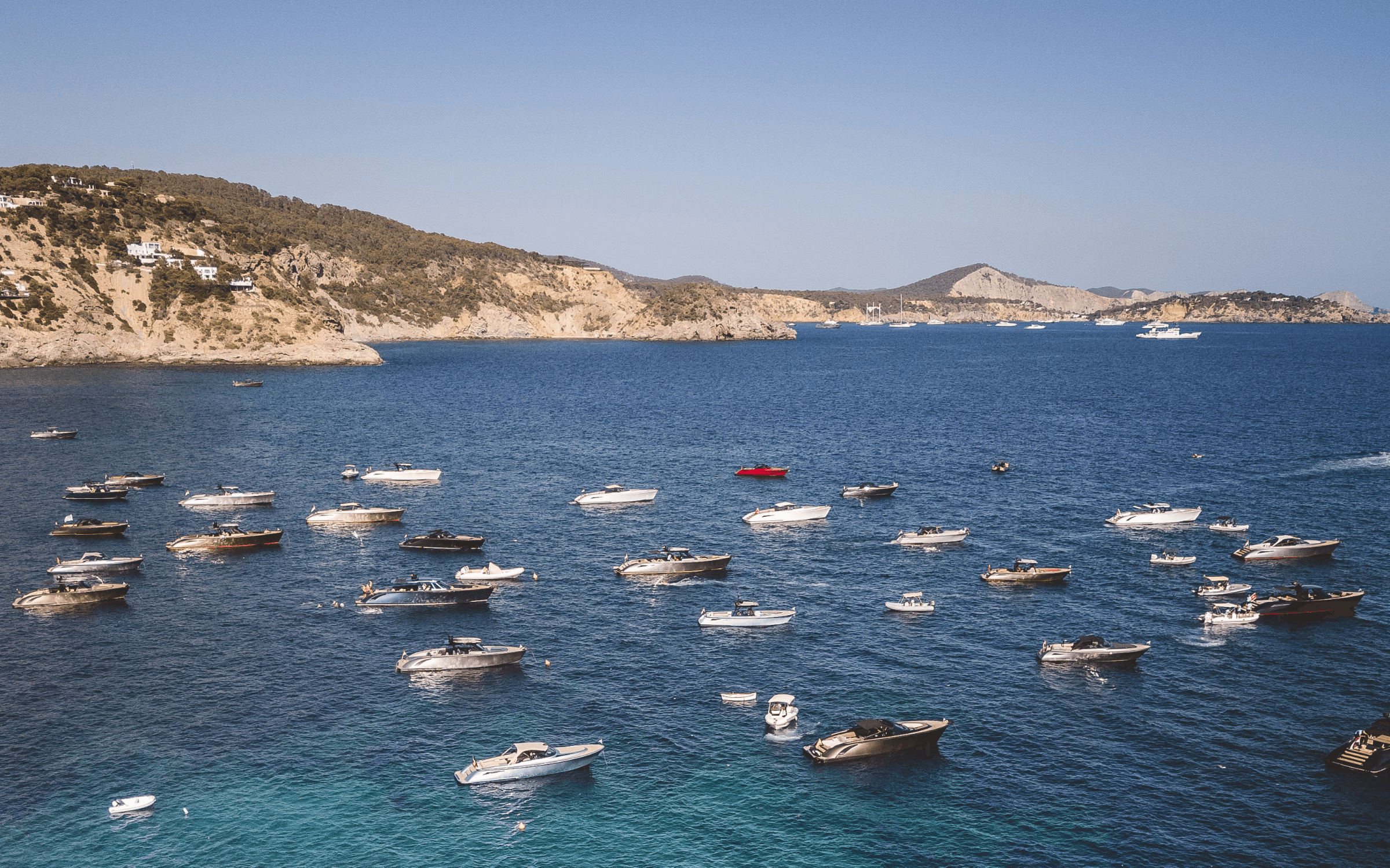 Numerous boats anchored in a calm bay with a scenic coastline and clear blue skies.