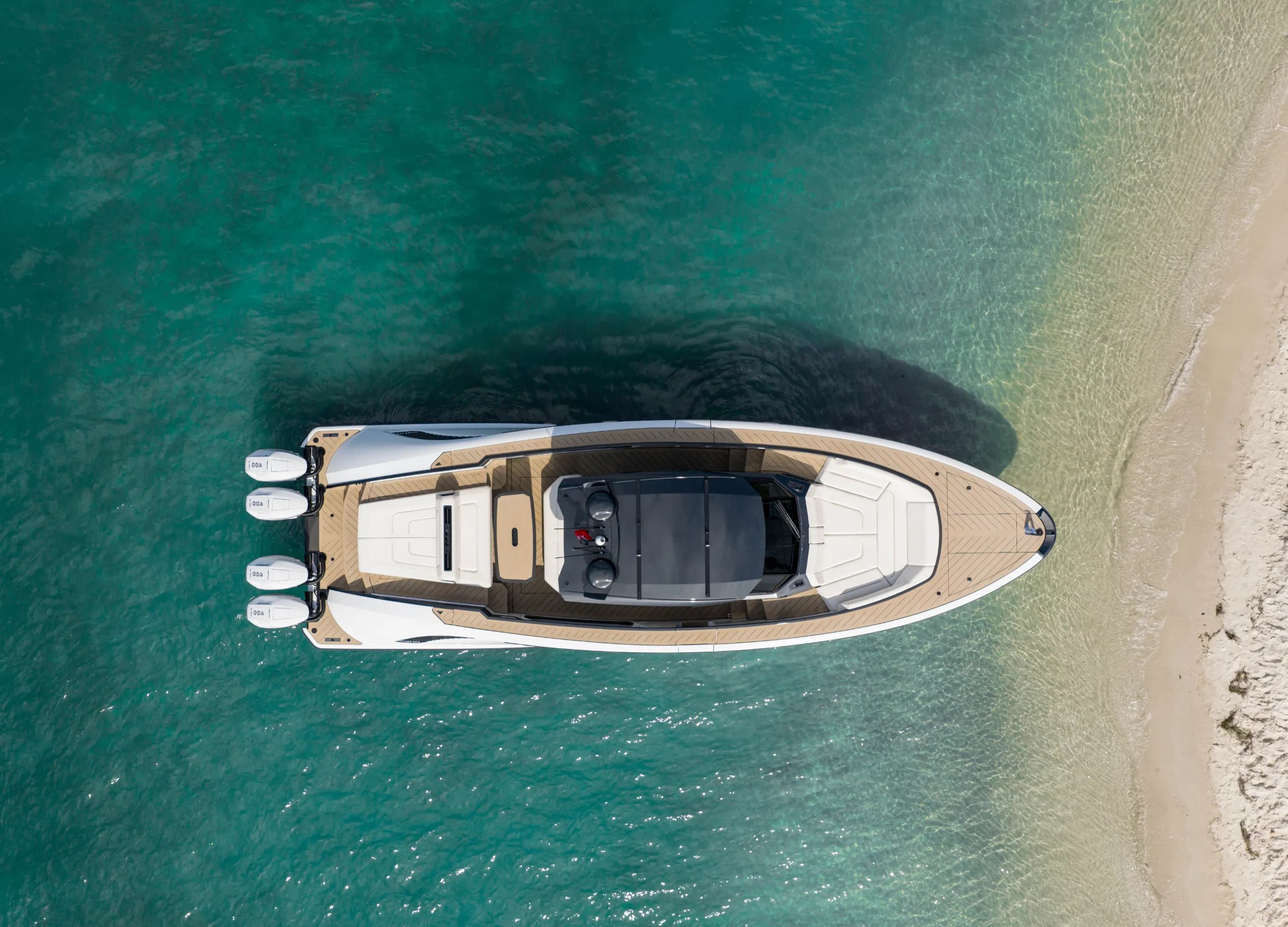 Aerial view of a sleek white speedboat with four outboard motors, floating near a sandy shore in clear turquoise water.