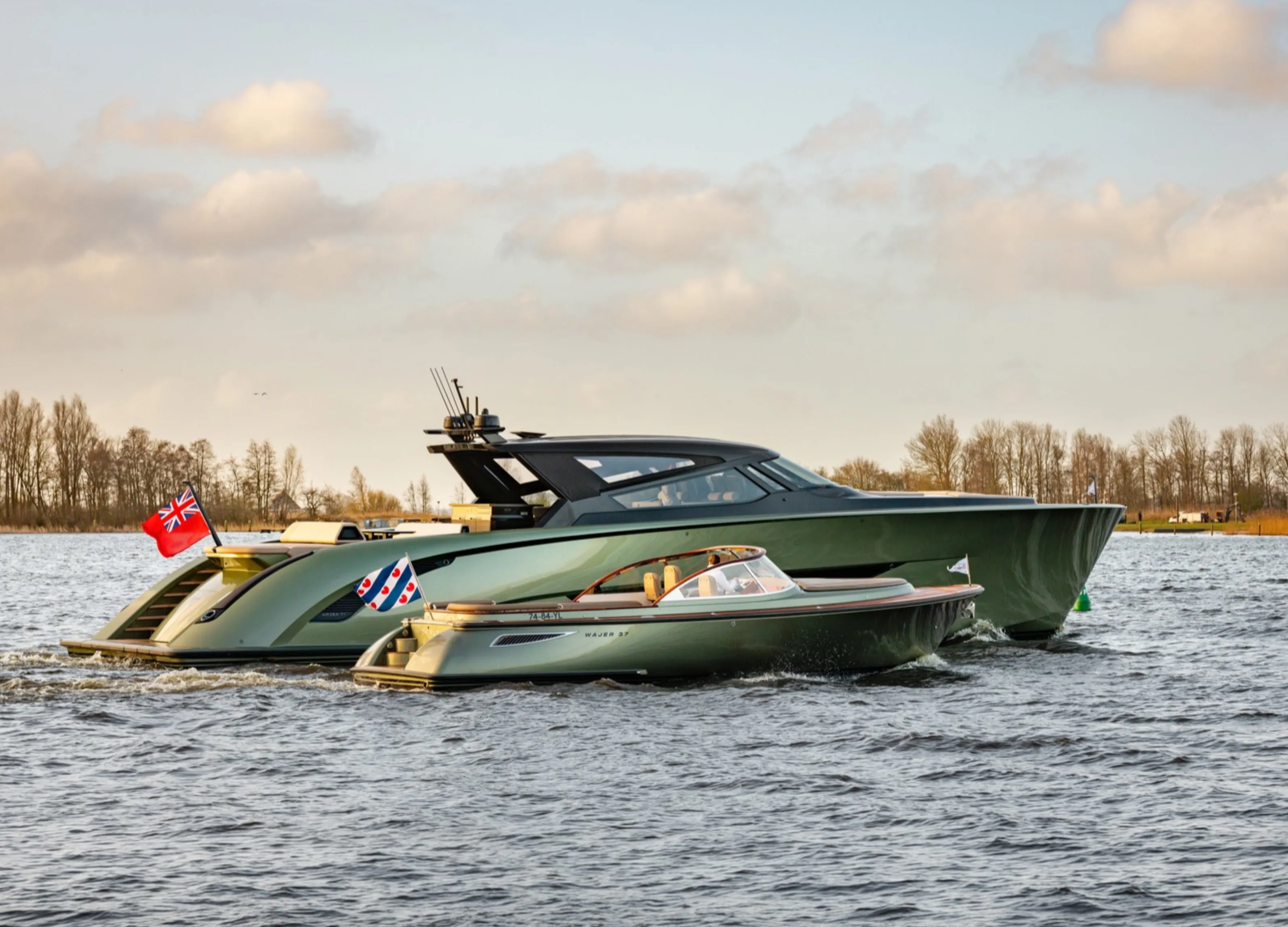 A large and a small modern yacht on a calm water body under a partly cloudy sky, with flags at the stern.