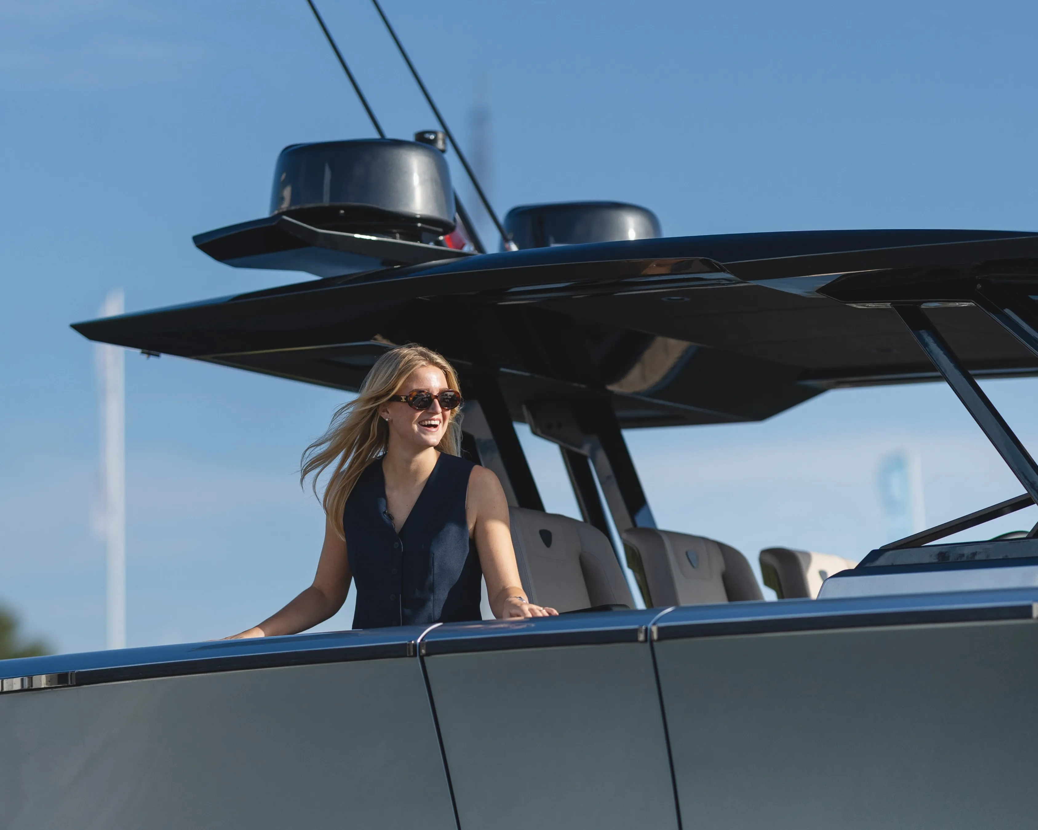 A woman with sunglasses stands smiling on a sleek, modern boat under a clear blue sky.