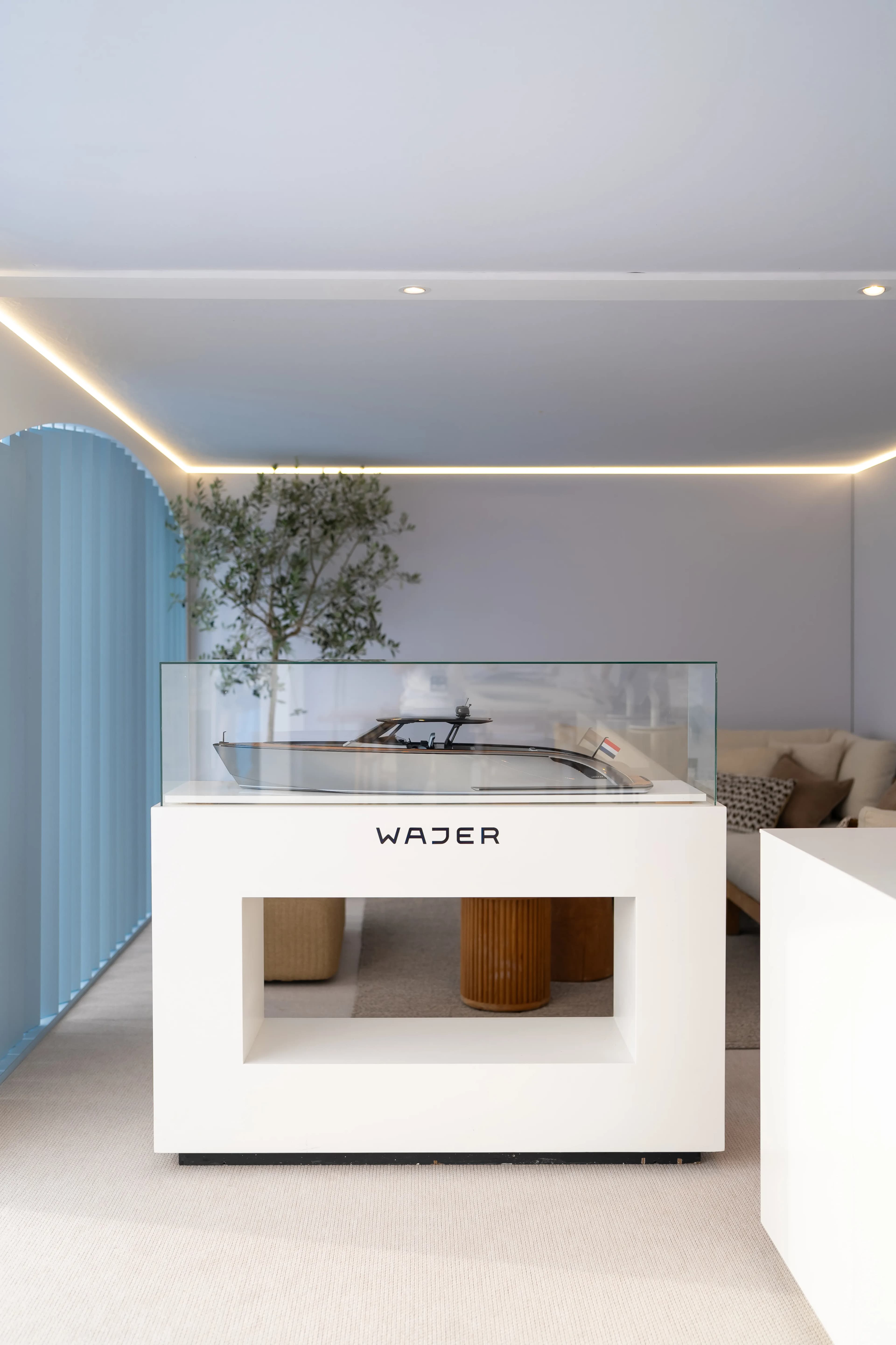 Modern interior showcasing a model yacht in a sleek, white display case with glass top and ambient lighting, a plant, and beige seating behind.