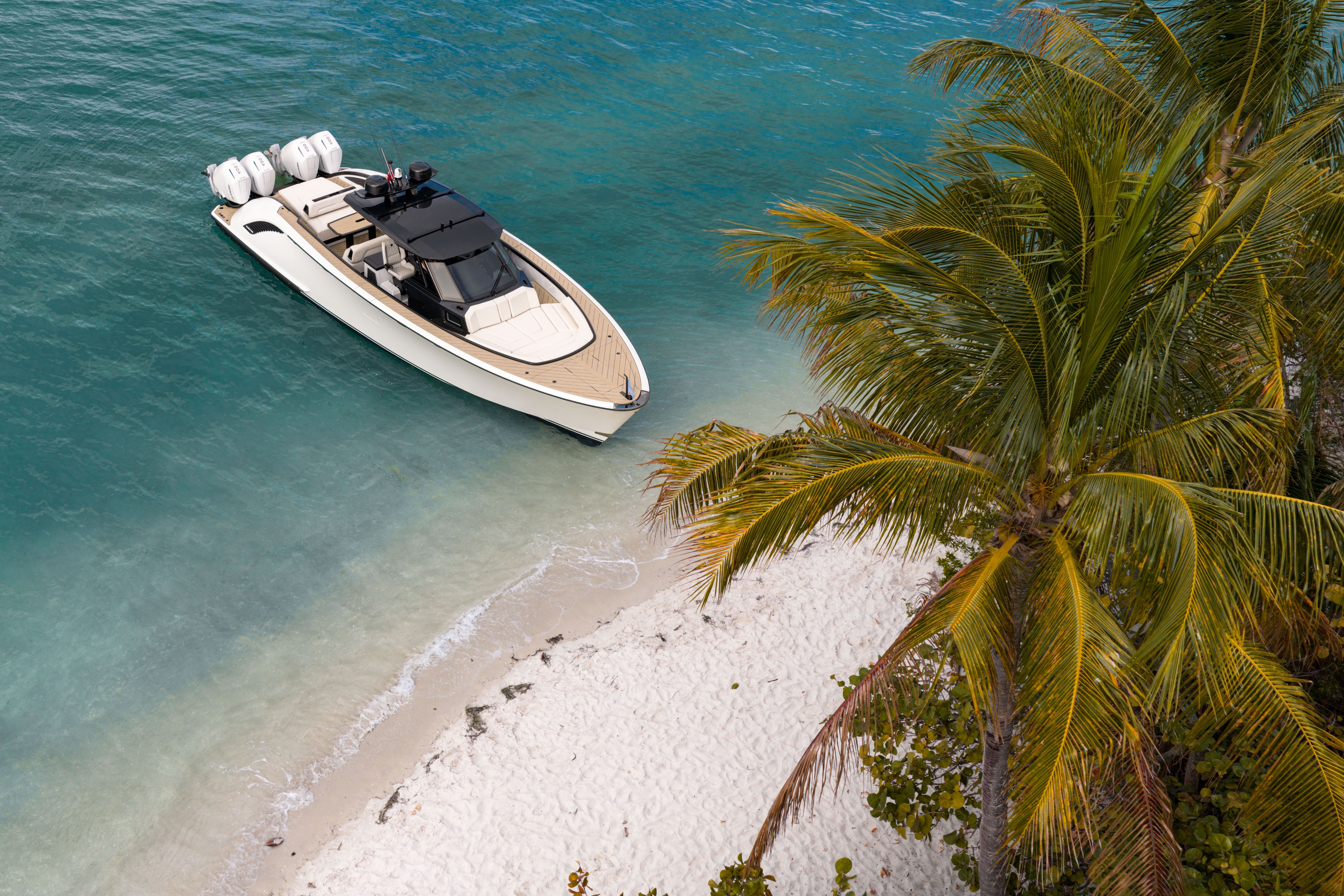 A sleek white speedboat with three outboard motors is moored near a sandy beach lined with palm trees, in clear turquoise water.